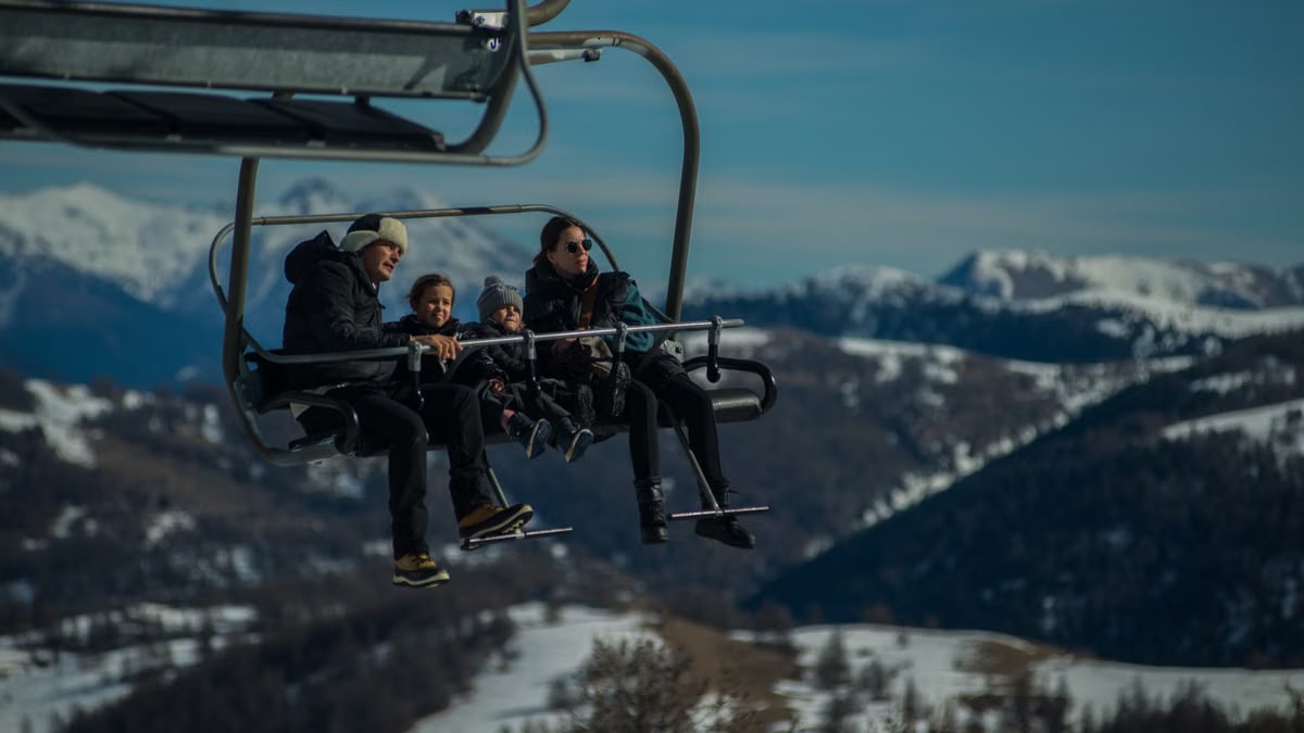 Family having fun on a ski lift with snow-covered mountains in the background during a sunny winter day.